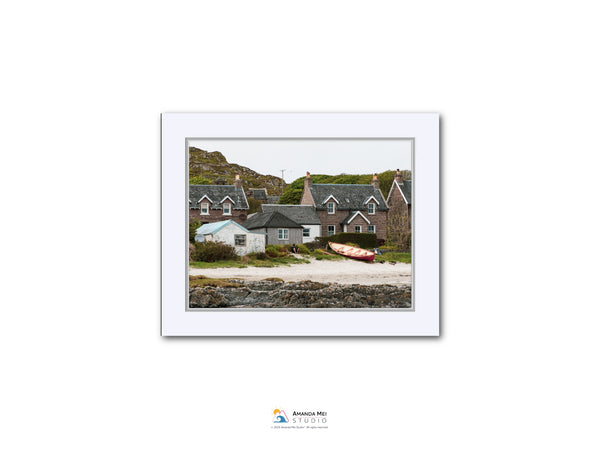 A white-framed photograph of a coastal village with houses, boats, and a rocky shoreline.