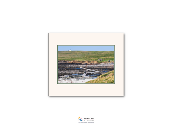 The image is a framed photograph depicting a scenic landscape with a winding path, a small white building, and a grassy hill in the background.