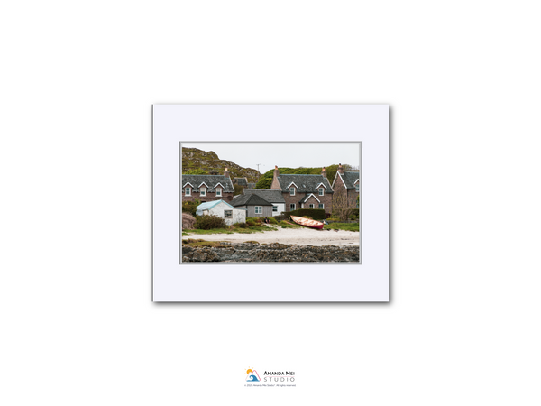 A white frame containing a photograph of a coastal village with houses, boats, and a rocky shoreline.