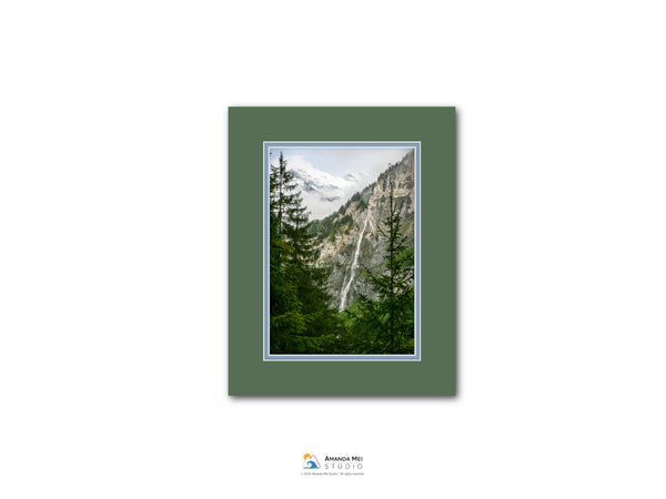 The image is a framed photograph of a mountainous landscape with a waterfall, surrounded by pine trees.
