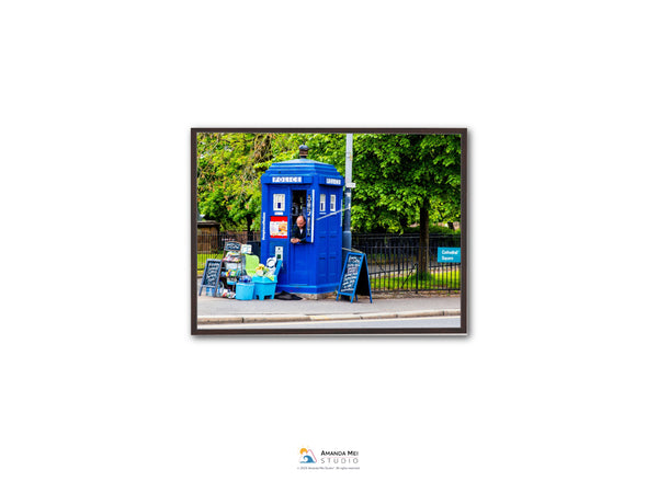 A blue telephone booth stands on a street, with a person inside the booth and various items displayed outside.