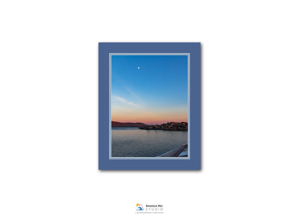 The image is a framed photograph of a serene body of water at sunset, with a small town or village visible on the opposite shore.