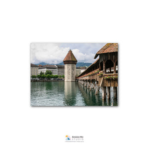 a picturesque scene of a wooden bridge with a tower on the left side, spanning across a body of water. The bridge is surrounded by lush greenery, and the sky above is filled with clouds, creating a serene atmosphere.