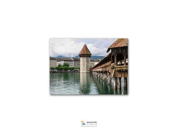 a picturesque scene of a wooden bridge with a tower on the left side, spanning across a body of water. The bridge is surrounded by lush greenery, and the sky above is filled with clouds, creating a serene atmosphere.