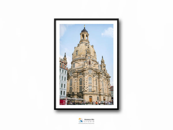 A color photo of a tan and gray baroque domed church on a blue partly cloudy day. There is a partial view of the building on the left and in the background on the right. This photo sits in a black thin frame with a white border.