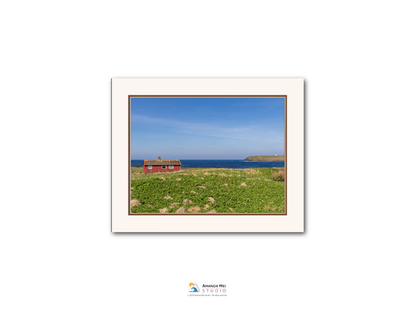 A red house sits on a grassy field, overlooking a body of water and a blue sky with clouds.