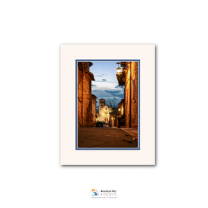 The image is a framed photograph of a narrow street in a European city at dusk, with illuminated buildings and a blue sky in the background.
