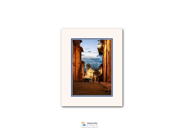 The image is a framed photograph of a narrow street in a European city at dusk, with illuminated buildings and a blue sky in the background.