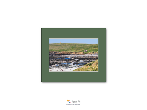 The image is a framed photograph of a coastal landscape, featuring a lighthouse on a hill, a rocky shoreline, and a body of water.