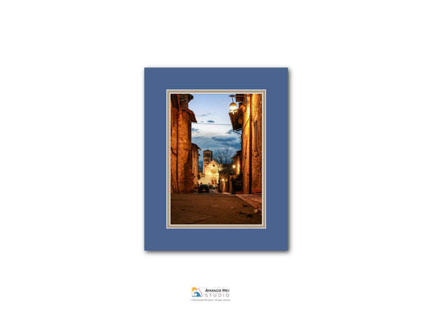 The image is a framed photograph of a narrow street in a European town at dusk, with illuminated buildings and a clear blue sky.