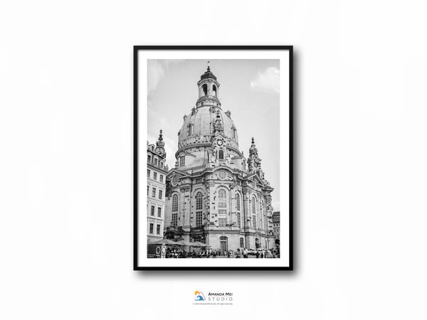 A black and white photo of a baroque domed church on a blue partly cloudy day. There is a partial view of the building on the left and in the background on the right. This photo sits in a black thin frame with a white border.