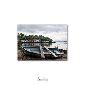 A small wooden boat rests on the shore of a body of water, with a row of colorful buildings visible in the background.