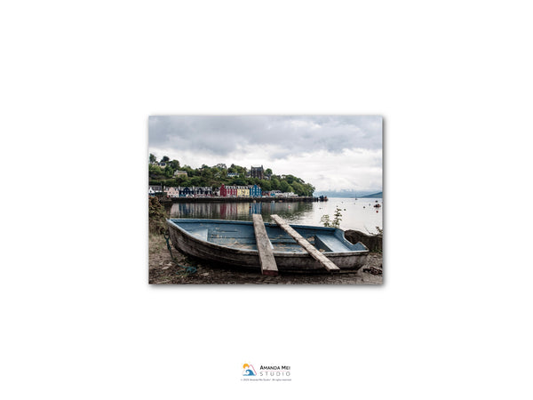 A small wooden boat rests on the shore of a body of water, with a row of colorful buildings visible in the background.
