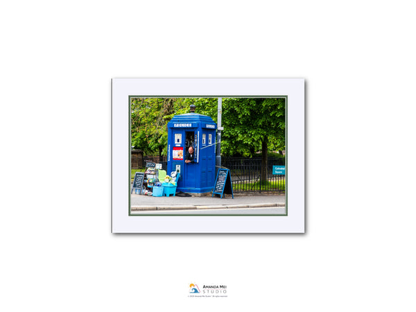 A blue telephone booth is situated on a street, surrounded by trees and a fence.