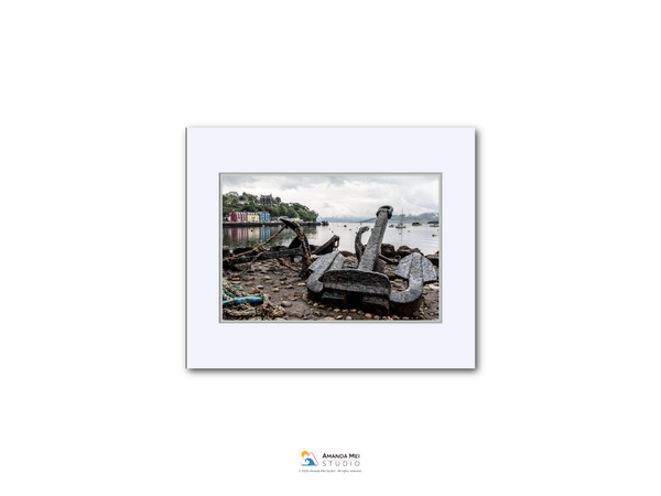 a large, rusted anchor on a beach, with a body of water and a building in the background.