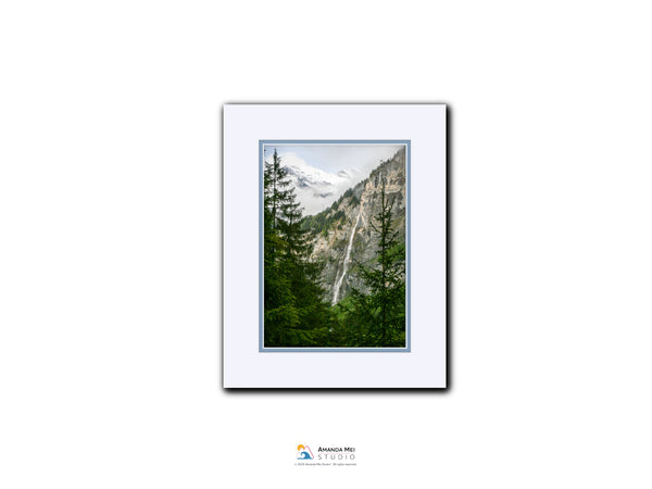 A framed photograph of a mountainous landscape with a waterfall, surrounded by pine trees.
