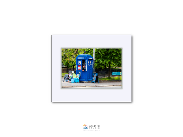 A blue telephone booth is situated on a street, surrounded by trees and a fence.