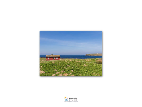 A red house sits on a grassy field, overlooking a body of water and a distant island.
