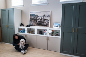 Amanda sits on the floor in a modern room, holding a small white dog. Behind her is a built-in white cabinet with glass-front shelves and beige lower cabinets, flanked by tall dark green wardrobes.