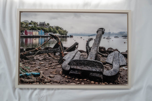 a rocky shoreline with a large anchor and several smaller anchors, as well as a body of water with boats in the distance.
