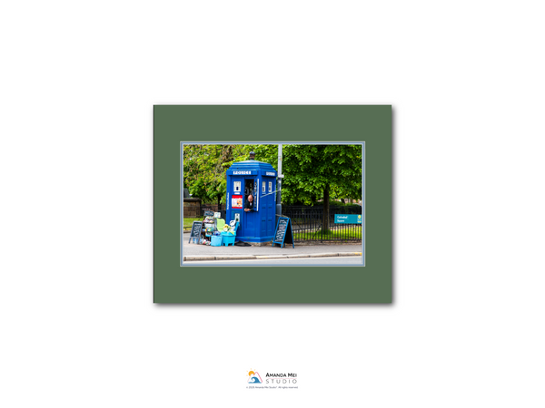 A blue telephone booth is situated on a street, surrounded by trees and a fence.