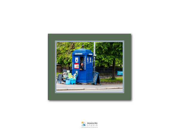 A blue portable phone booth is situated on a street, surrounded by trees and a fence.