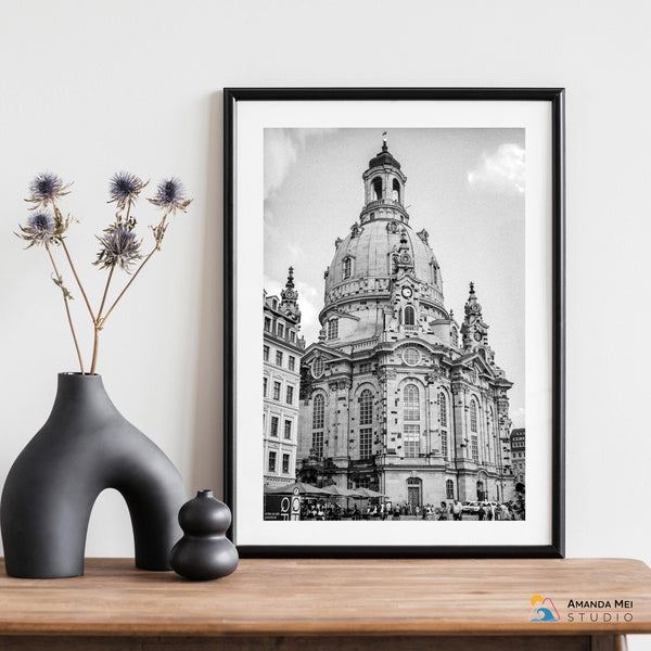 This is a table top with a black vase with some dried flowers next to a smaller black vase. Next to that, there is a small black and white print in a black frame.