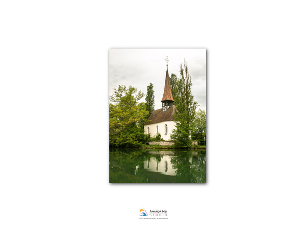A white church with a steeple sits on the shore of a calm lake, surrounded by lush greenery.