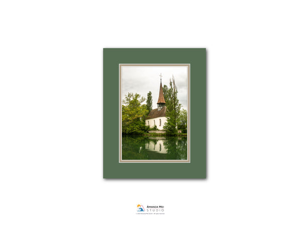 A framed photograph of a white church with a steeple, surrounded by trees and reflected in a body of water.