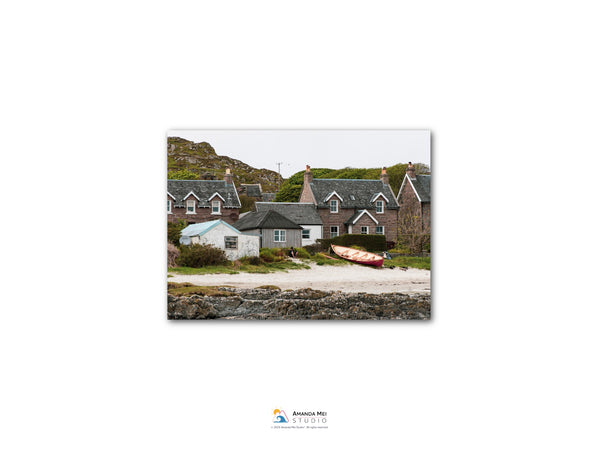 A coastal village with houses and boats on the beach, set against a backdrop of rocky cliffs and a cloudy sky.