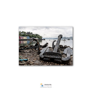 a rusty anchor on a rocky shore, with a body of water and buildings in the background.