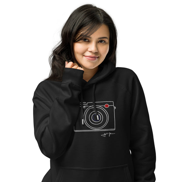 A young woman with dark hair is smiling and posing for the camera, wearing a black hoodie with a white graphic of a camera on it.