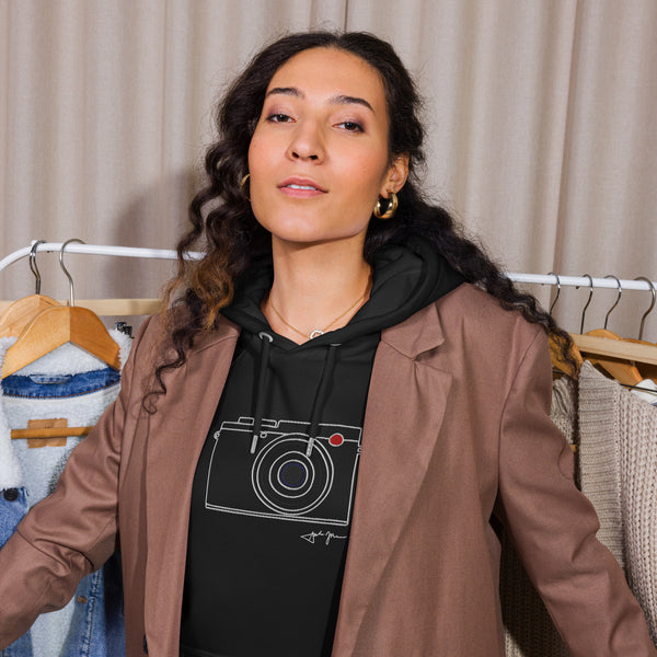 A woman with curly hair stands in front of clothing racks, wearing a brown jacket and a black hoodie with a camera graphic.